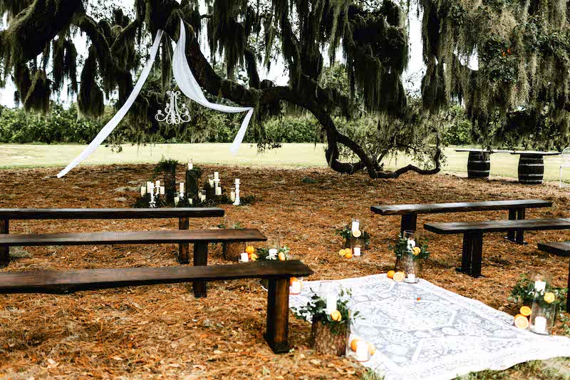 dark wood benches set up for outdoor wedding ceremony, with candles, flowers, and a carpet used as decoration