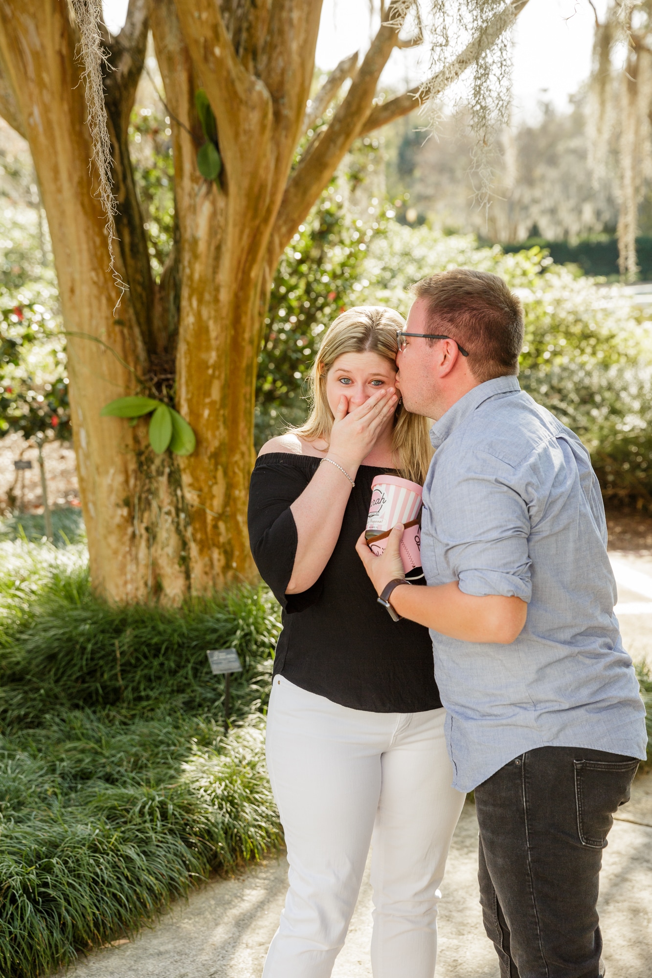 woman holds hand over mouth in shock while holding kelly's homemade ice cream marriage proposal container with custom proposal on it while man kisses her on the cheek