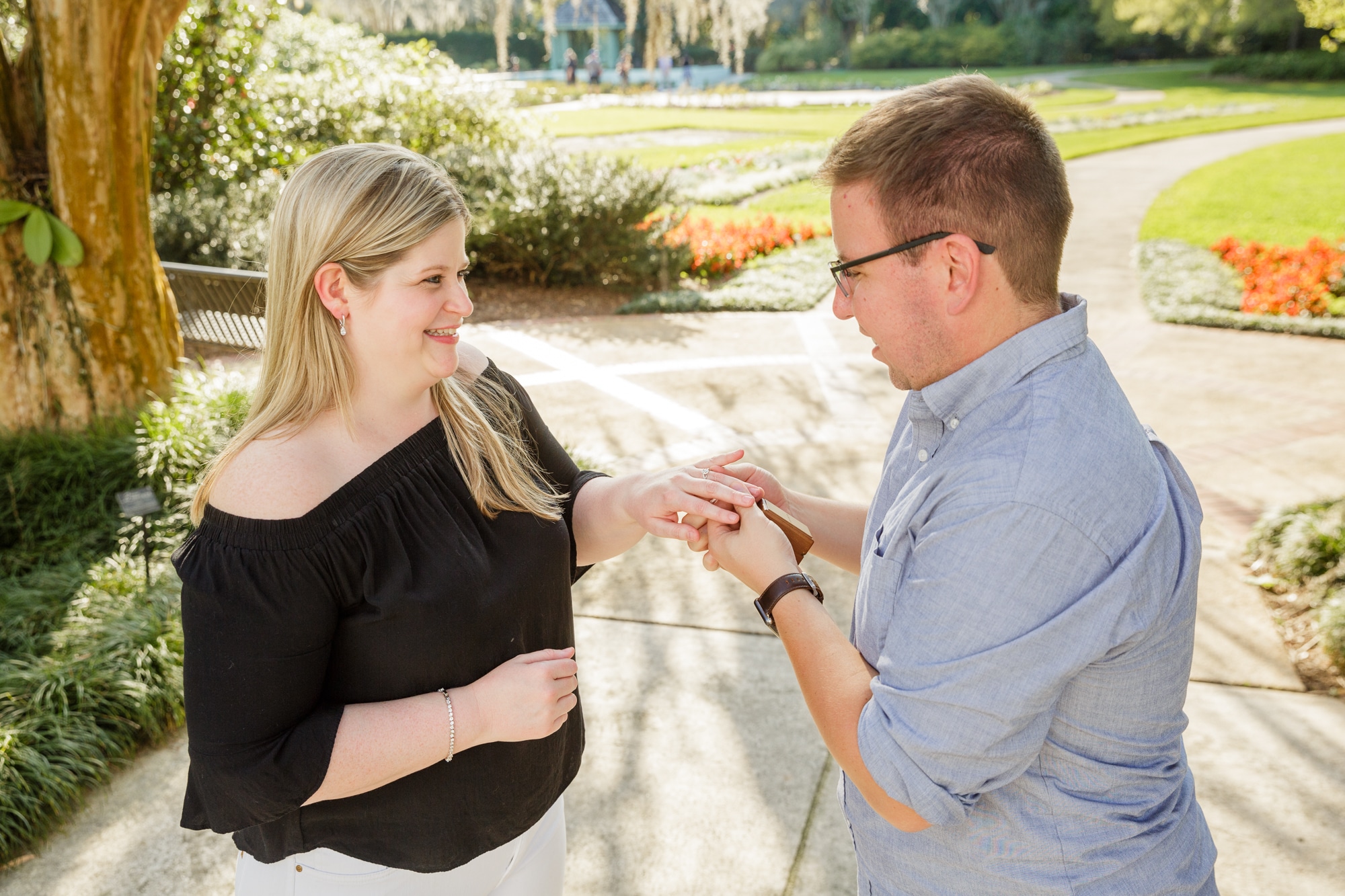 engaged couple stand outside while girl smiles and man puts the engagement ring on her finger