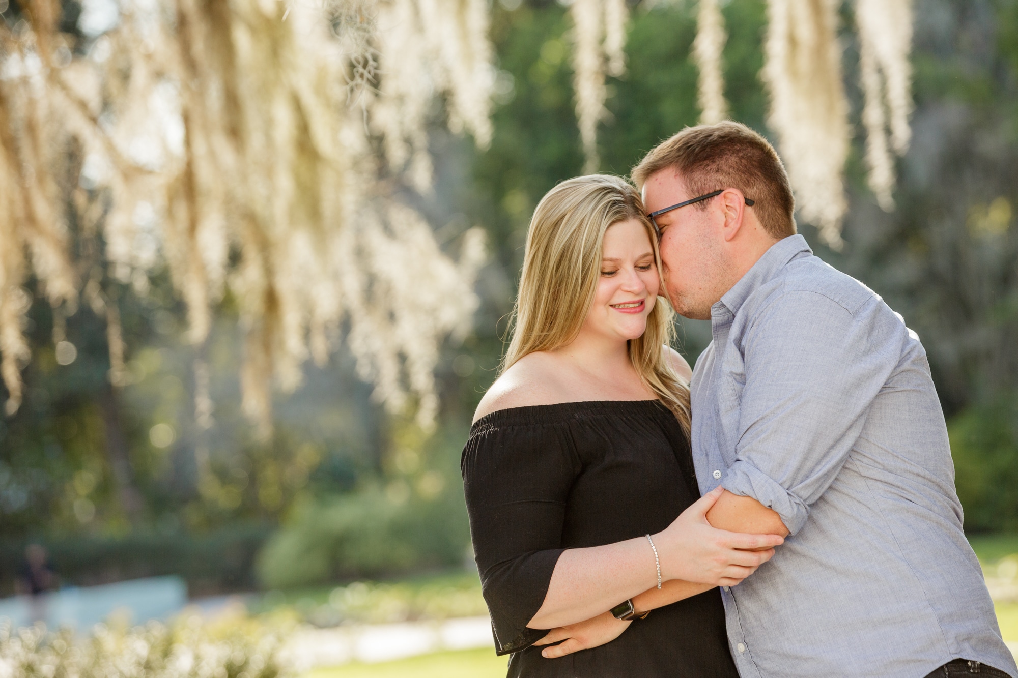 woman stands with off the shoulder black shirt on with eyes closed smiling as man leans over and kisses her on the cheek outside under tree with moss