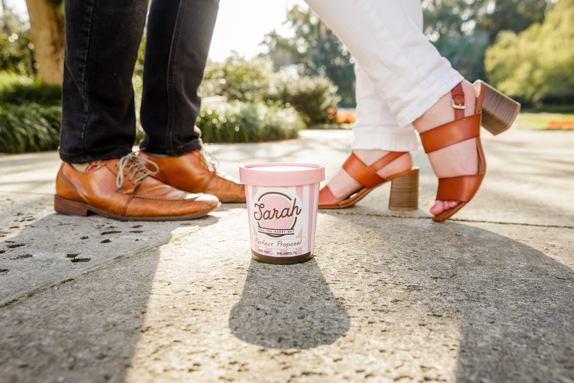 close up picture of couples feet with shoes on outside and a homemade proposal prop of a kelly's homemade ice cream container