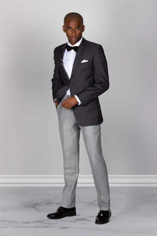 man wearing a dark grey suit jacket, light grey dress pants, black shoes, black bow tie, and a white pocket square