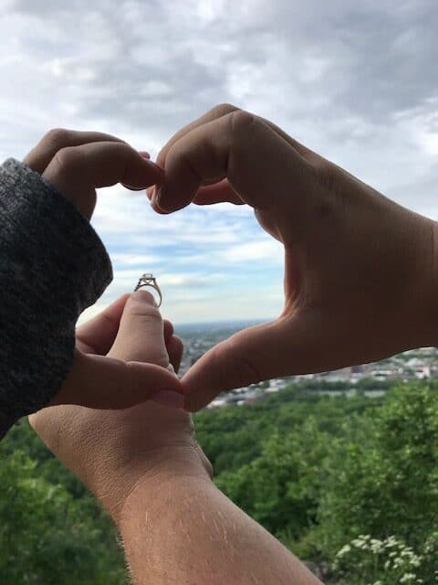 two different hands in camera view make up a heart in the middle with another arm outstretched in front holding an engagement ring with the sky and trees in the background