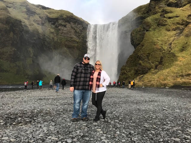 man wearing plaid shirt and woman wearing a scarf stand next to each other in front of huge waterfall surrounded by greenery