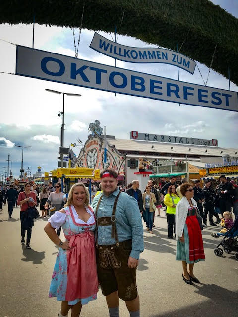 couple stands together for picture wearing traditional german clothing under a sign that read oktoberfest with people in the background