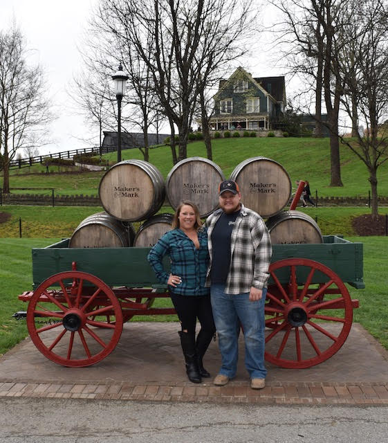 couple stands in front of giant wagon with red wooden wheels holding 6 stacked wooden barrels outside