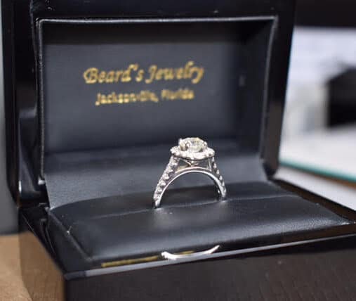 side view of an engagement ring sitting in a black ring box from a local florida jewelers name at the top of the box in gold writing