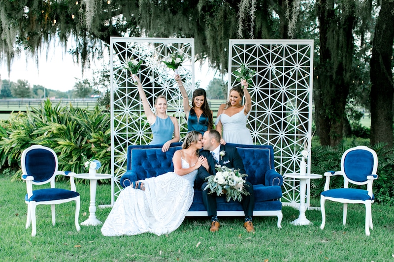bride and groom kissing on a blue couch while three bridesmaids hold their flower bouquets up behind them.