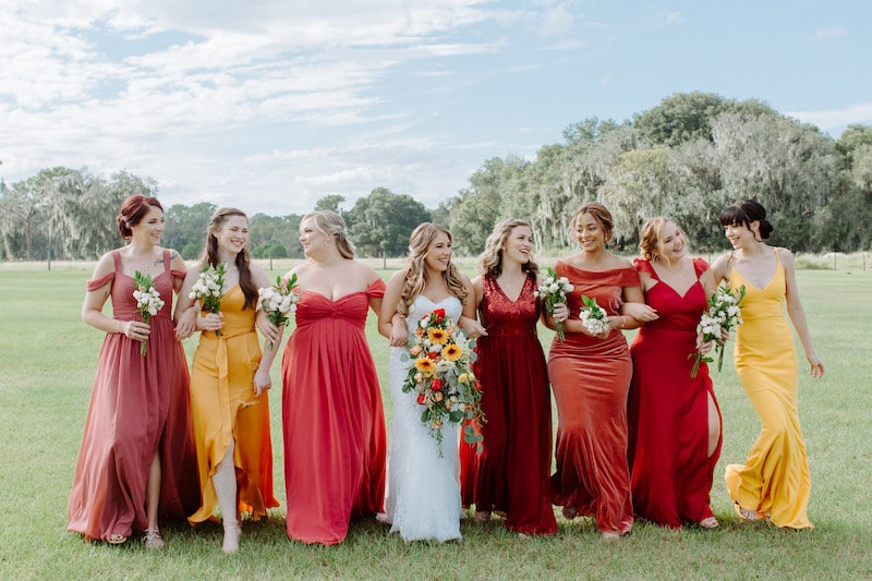 bride standing in the middle of all of her bridesmaids who are wearing a dresses in a variety of reds and yellows