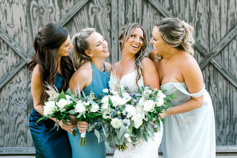 bride and her bridesmaids in shades of blue laughing while holding their matching flower bouquets