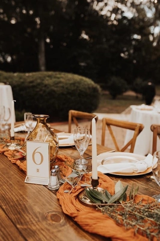 long wooden tables set up for wedding reception with orange linen runner, candles, and matching table number