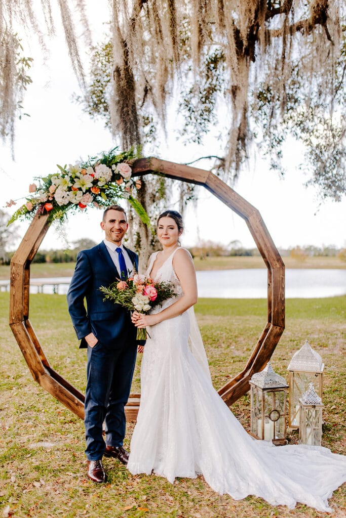 bride and groom standing in front of wood circle with floral garland under oak trees at legacy at oak meadows