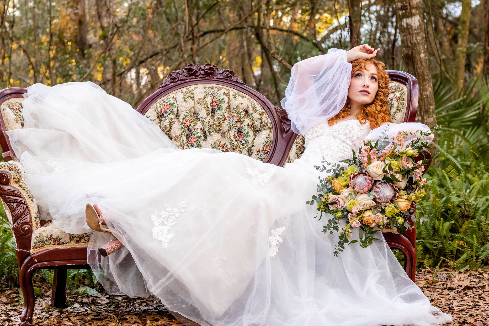 bride in fluffy wedding dress laying on vintage floral couch outside in woodsy area with hand on head and bouquet beside her