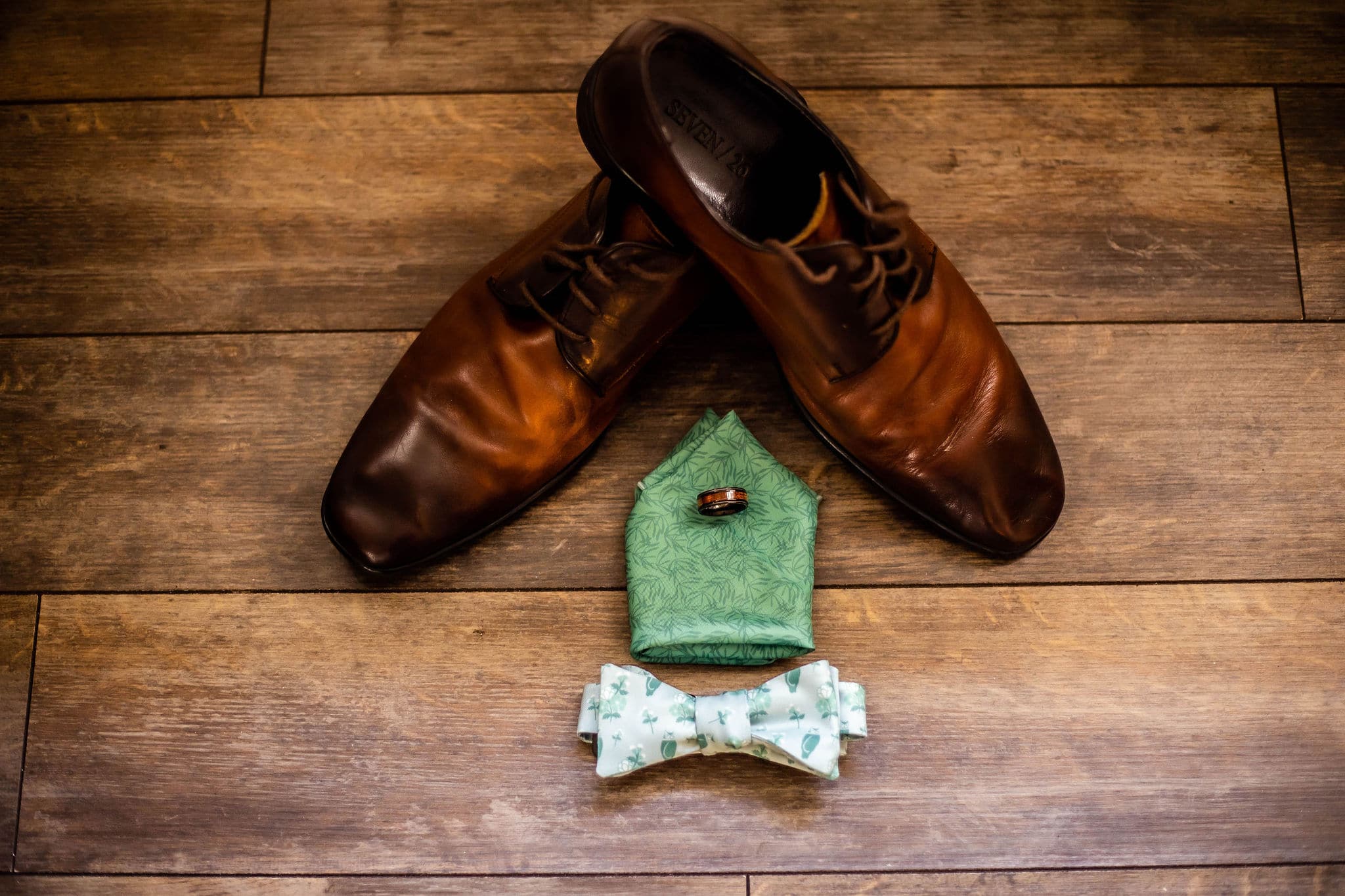 detail shot of grooms attire for wedding day with brown dress shoes, green patterned pocket square with ring sitting on it and bowtie in front of that on a wood plank flooring