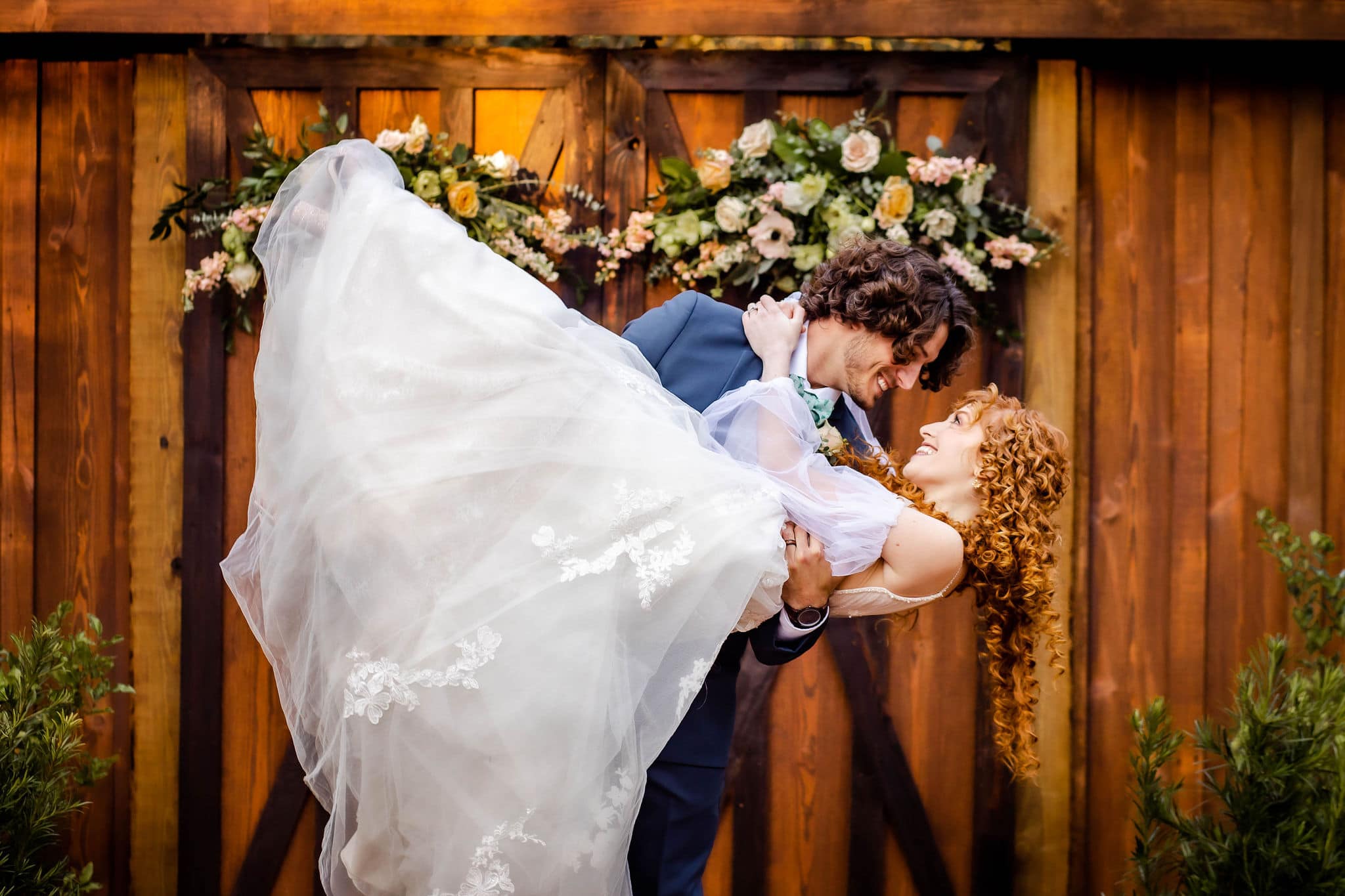 bride being dipped down by groom outside in front of barn doors with hanging floral arrangements attached to them