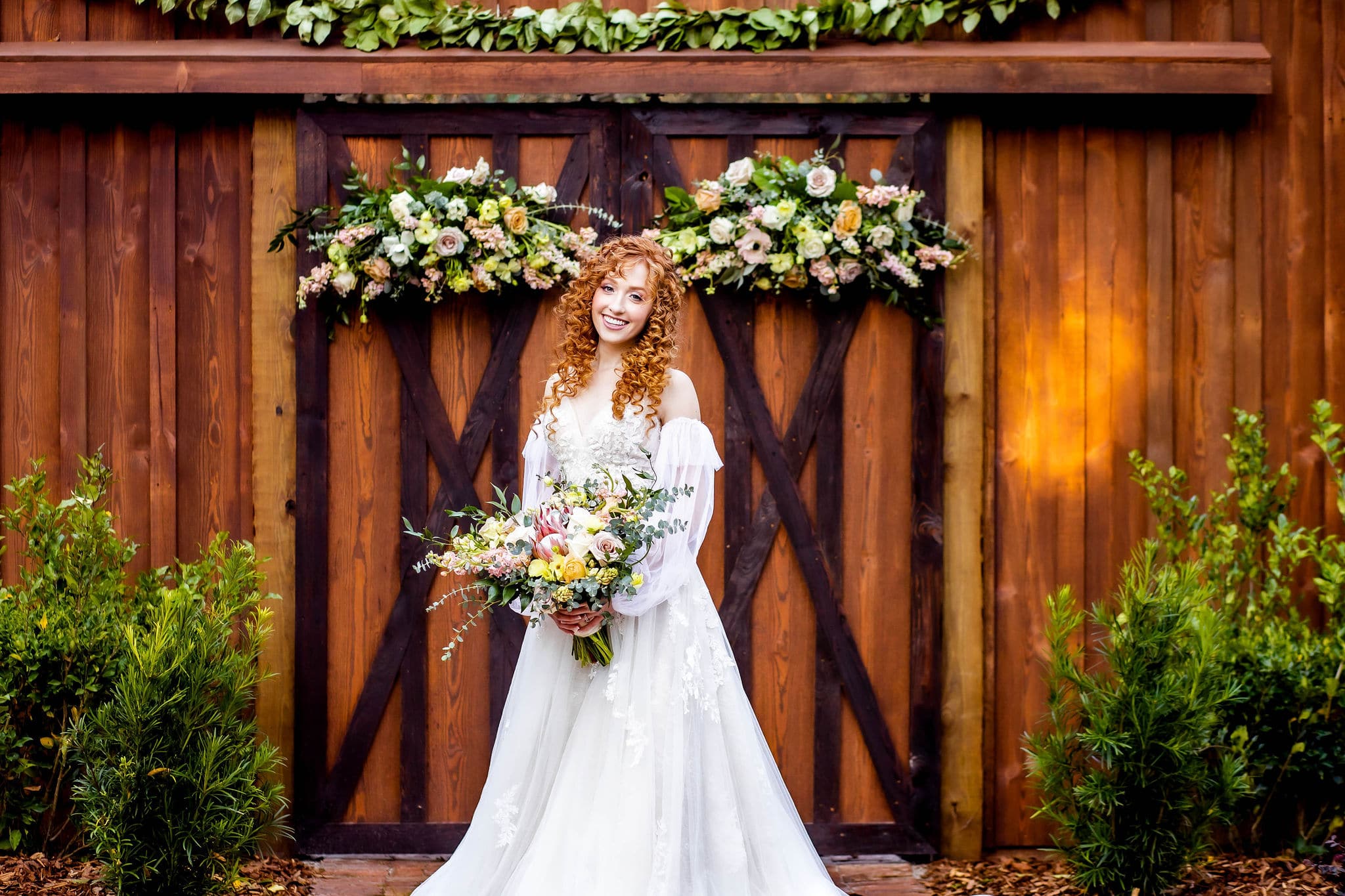 woman in bridal gown holding bouquet stands in front of closed barn doors with greenery in front of them off to the sides and two floral arrangements hanging from the top of them