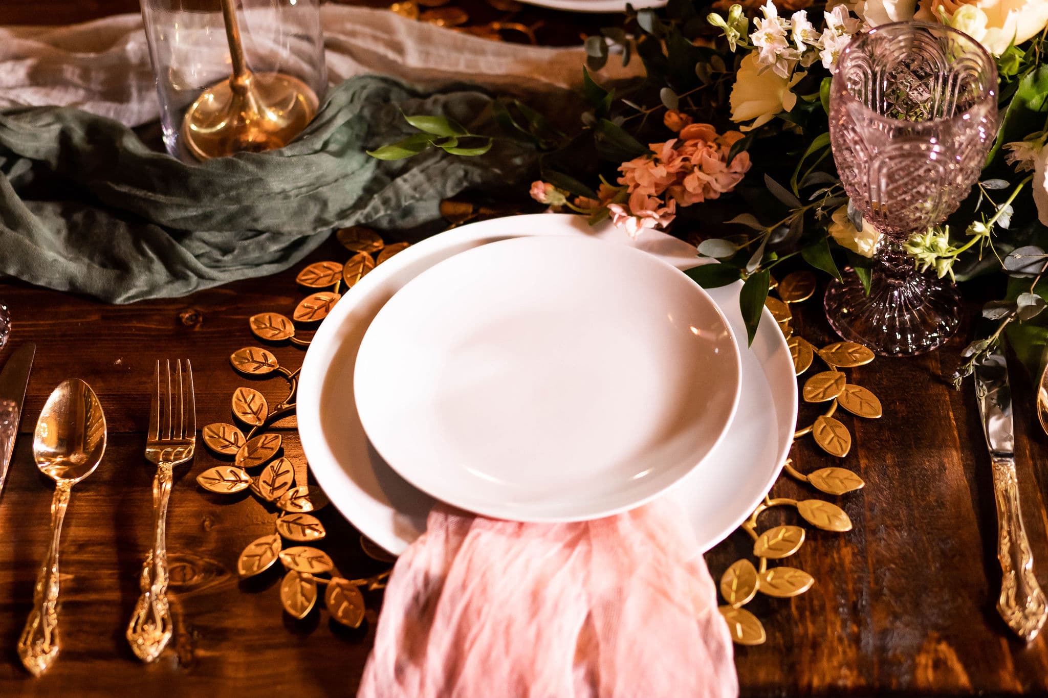 place setting on wedding day reception able with gold leaf charger and white plates whit pink napkin flowing from under them with gold flatware on natural wood table