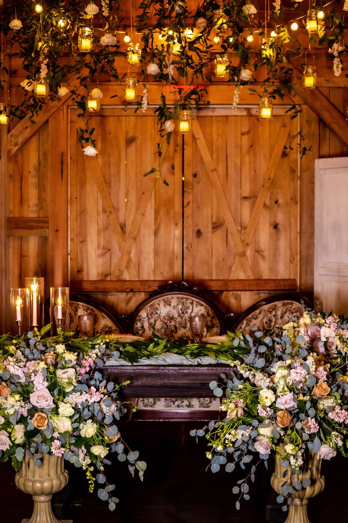 sweetheart table at wedding reception decorate with huge floral arrangements on the floor in front and vintage floral couch for seating with candlesticks on the table and hanging lights with greenery above it