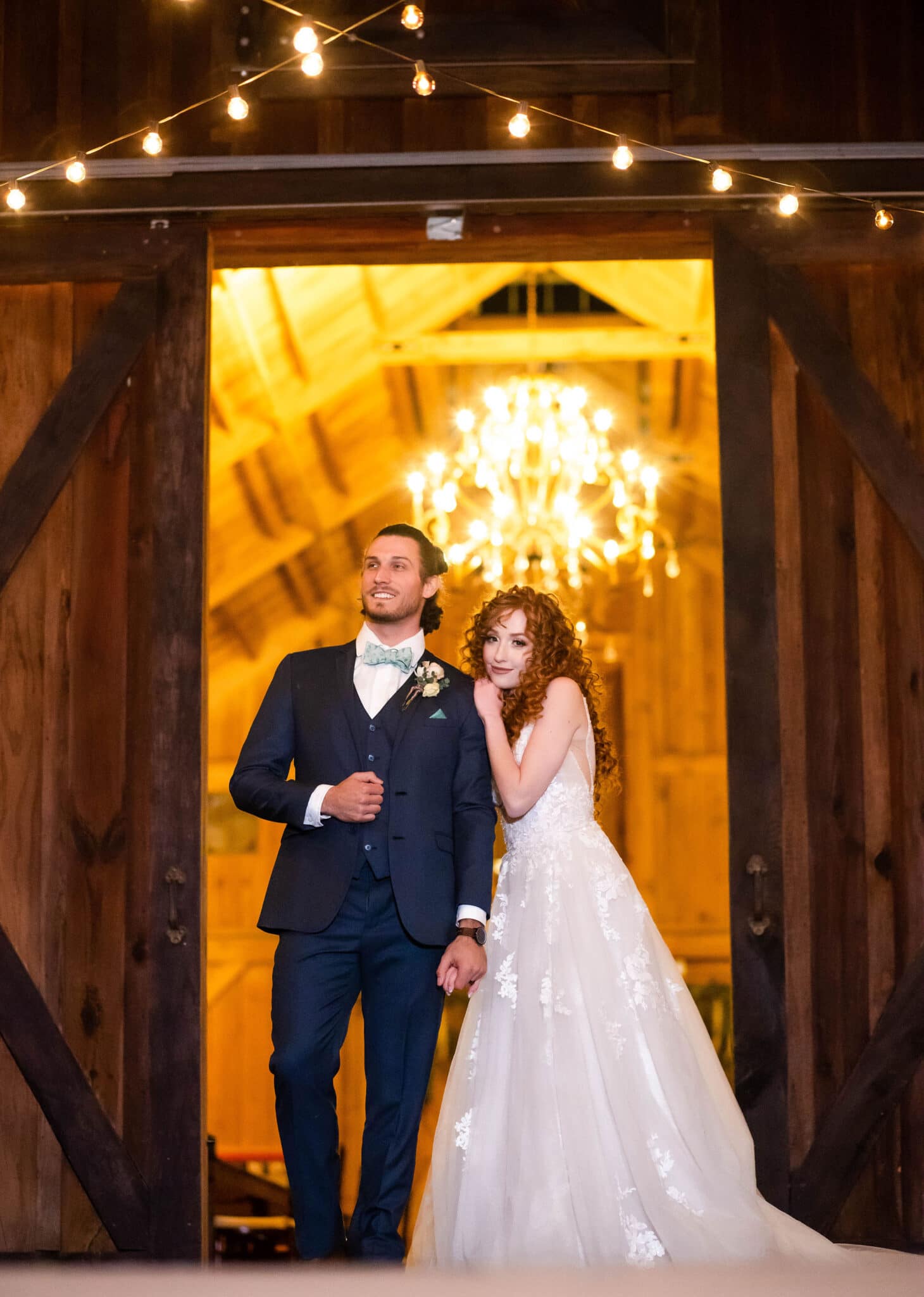 married couple at wedding outside of barn reception space looking off into the distance with big chandelier hanging inside behind them