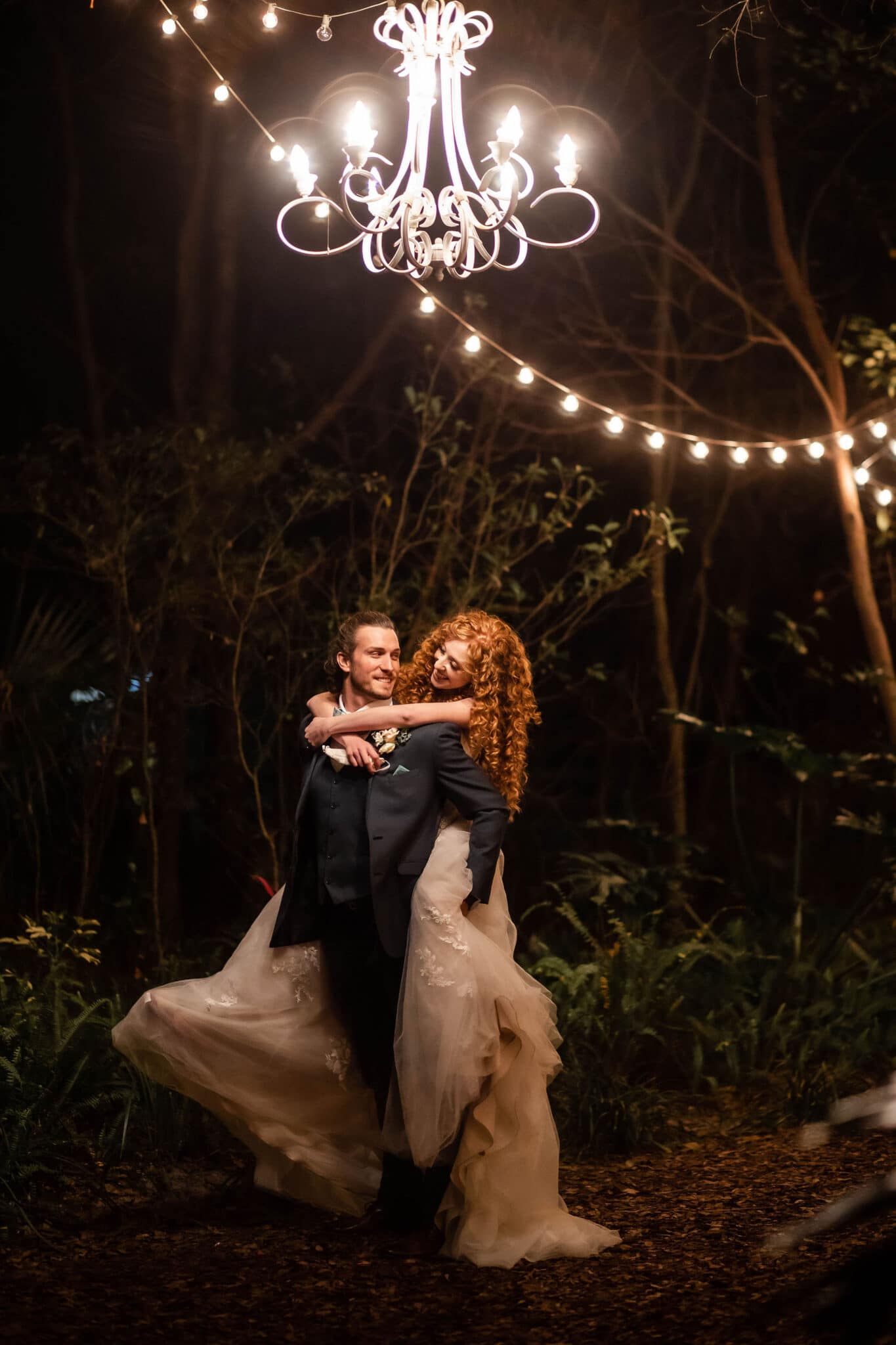 groom carries bride on his back as she looks at him outside at the night under chandelier hanging from trees