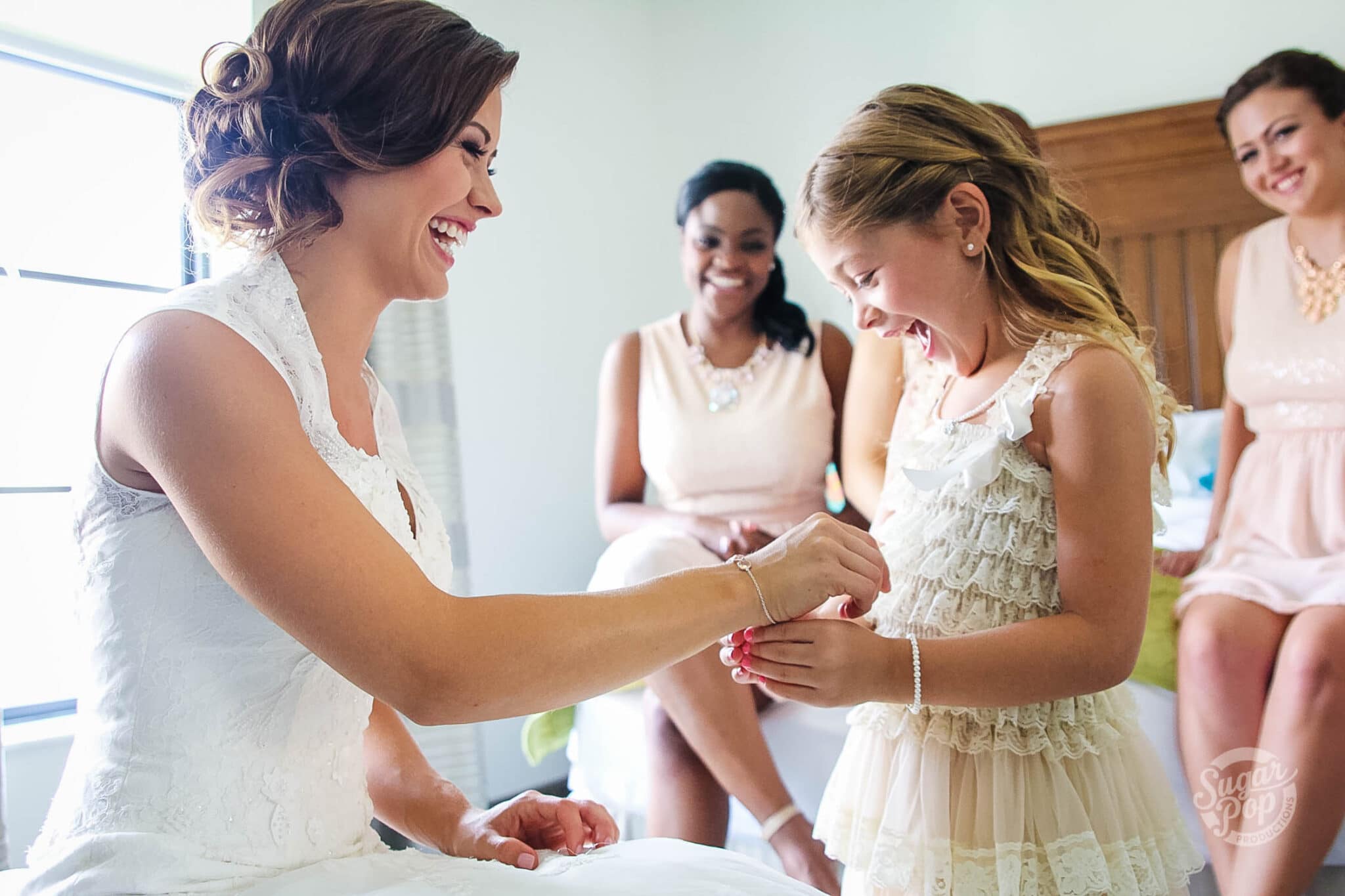 laughing bride lets little girl with ruffle dress on put a bracelet on her right wrist with two women in the background sitting on a bed