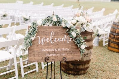 brown welcome sign for wedding ceremony with white and green floral garland in front of large barrels