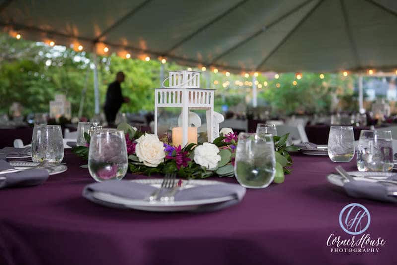 large tent set up for wedding reception, with tables that have purple tablecloths, matching flowers, and candlelit lantern