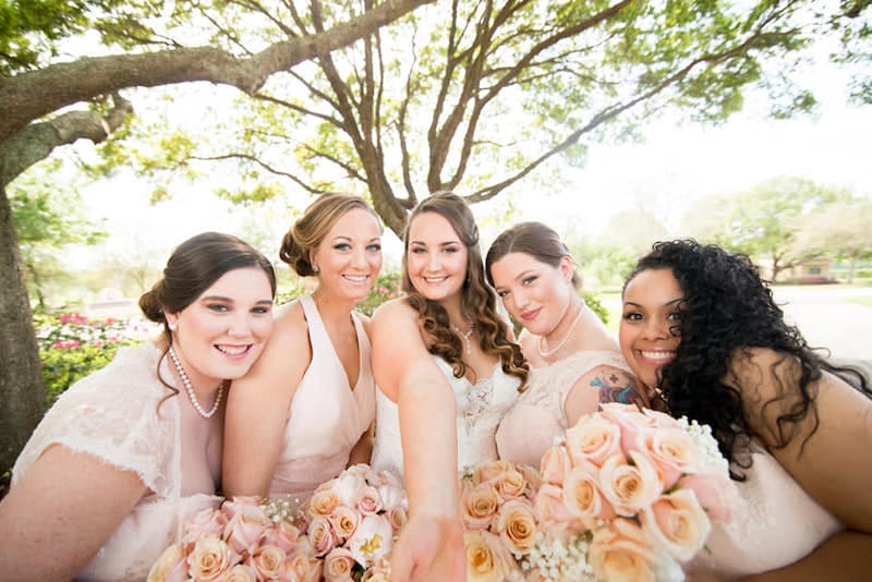 bride and her bridesmaids in matching dresses standing under a large tree and holding their matching flower bouquets