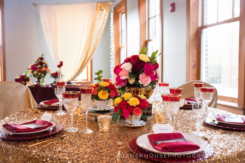 table decorated for wedding reception with flower centerpieces, a gold tablecloth, red napkins, and matching silverware and candle holders