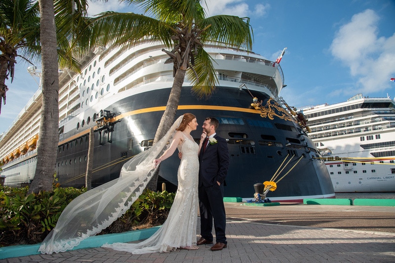 bride and groom standing beneath palm trees with a large Disney cruise ship behind them