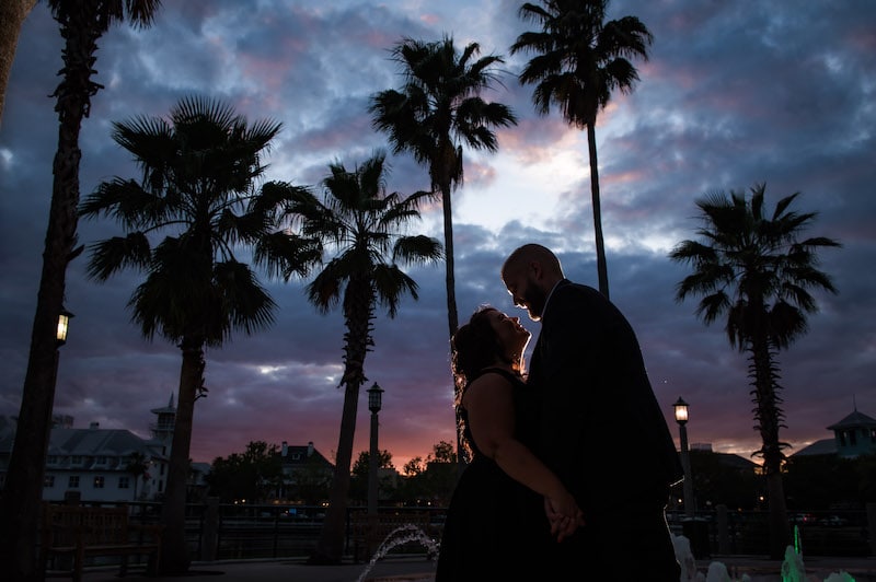 bride and groom kissing next to a water fountain at night with the sun nearly set behind them