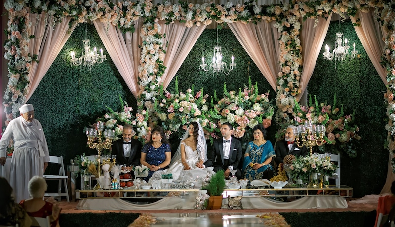 bride and groom sitting next to their parents with large flower displays covering the room