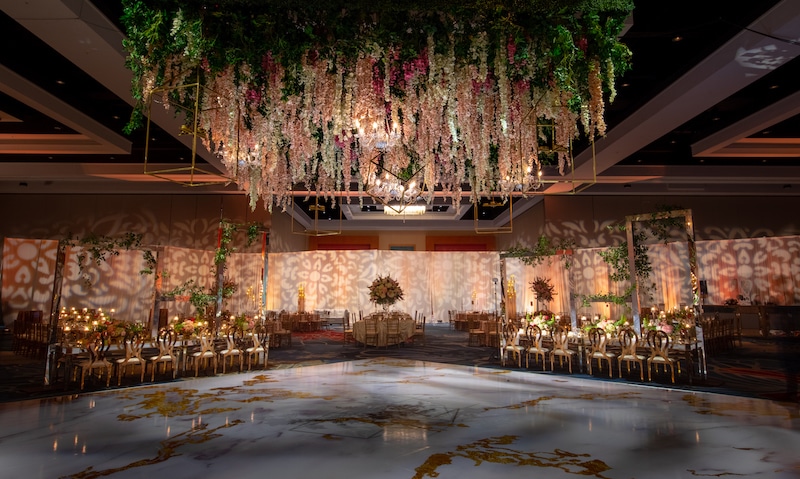 Eventrics Weddings preps indoor wedding reception area with flowers hanging from the ceiling, decorative lights along the wall, and candles on all the tables