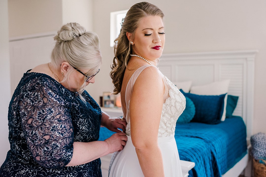 brides mother wearing navy blue dress helps bride zip dress up inside in front of bed with navy blue blanket