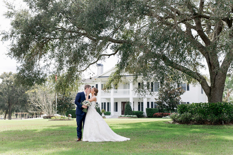 bride and groom kissing while standing in the shade of a large tree in front of beautiful white house