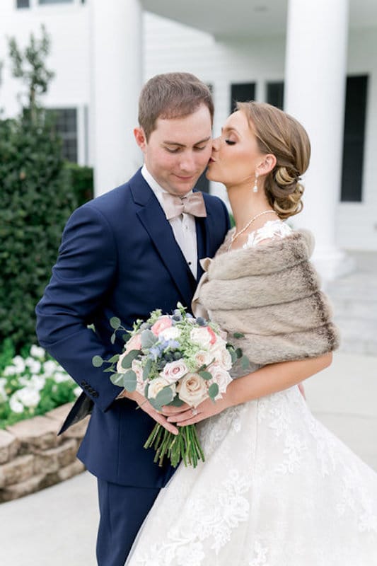 bride with winter stole on kissing her groom on the cheek while he looks down at her flower bouquet at event planned by Just Save the Date