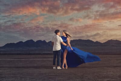 woman in a blue dress floating in the wind standing next to a man while in the middle of the desert with mountains behind them