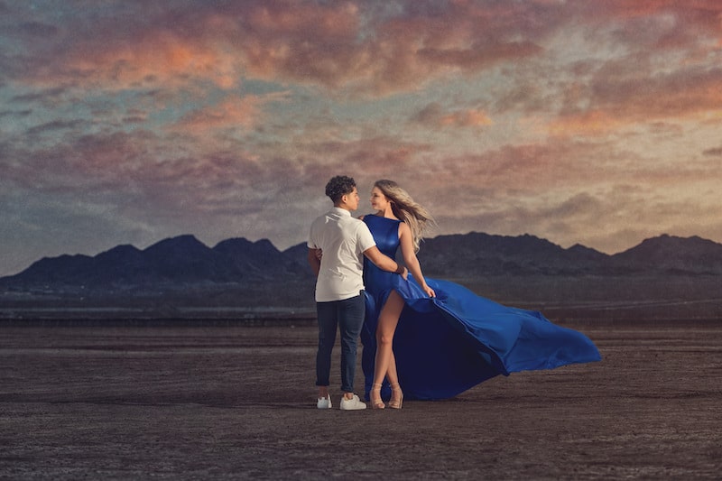 woman in a blue dress floating in the wind standing next to a man while in the middle of the desert with mountains behind them