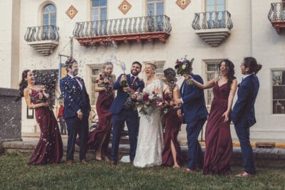 bride and groom plus bridal party laughing while groom pops a bottle of champagne