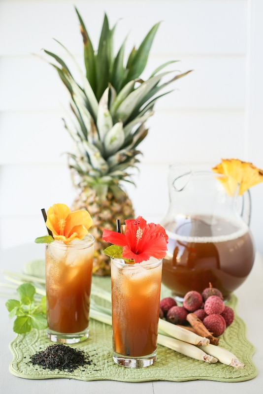 glasses filled with tropical drinks with flower garnishes on a green mat with a pitcher of iced tea, a pineapple, and more
