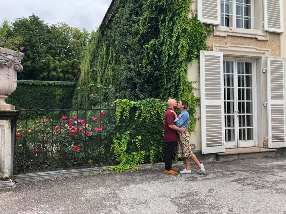 couple stand in front of building with a huge amount of greenery next to it kissing