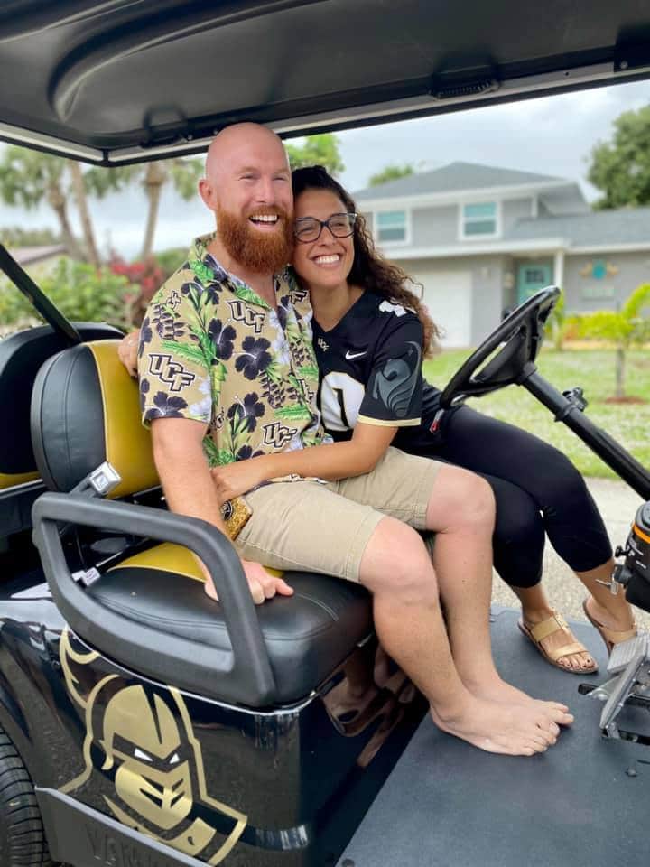 man wearing floral shirt and woman wearing football jersey sit on golf cart together with gold UCF knights logo on the side