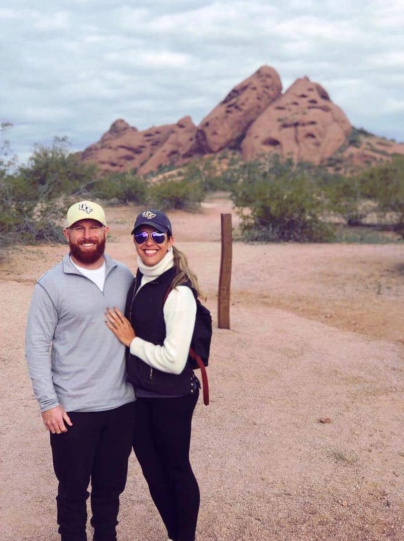 man and woman wearing long sleeves and pants with ball caps on stand in desert outside in front of mountain