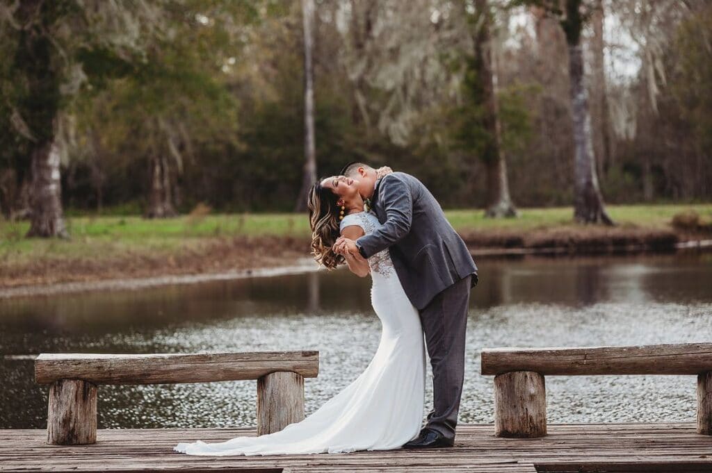 groom kissing his bride on her neck while both stand on a dock sticking out into a small lake