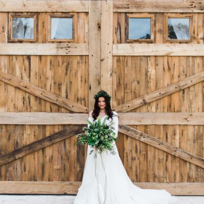 bride with floral crown holding large greenery bouquet in front of rustic wooden barn doors