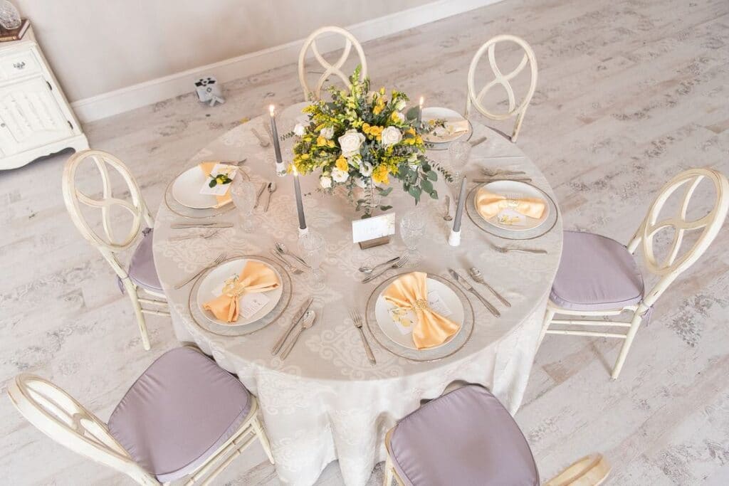 tables set up for wedding reception with purple chair cushions, silver candles, and peach napkins, and a white and yellow flower centerpiece