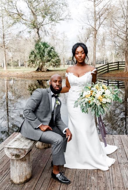 groom sitting on a wooden bench on a dock over a small body of water at Cypress Creek Farmhouse, with his new bride standing next to him