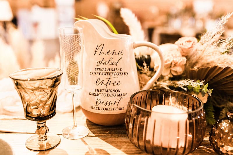 menu written in calligraphy by Bare Lettered Designs on the side of a water pitcher sitting on wedding reception table