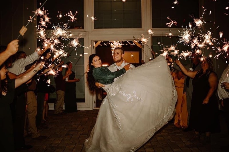groom carrying his bride while their guests hold sparklers at a wedding coordinated by Hearts and Souls Event Management