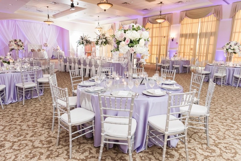large room decorated for wedding reception with purple linens and uplights with white and purple flowers for decorations